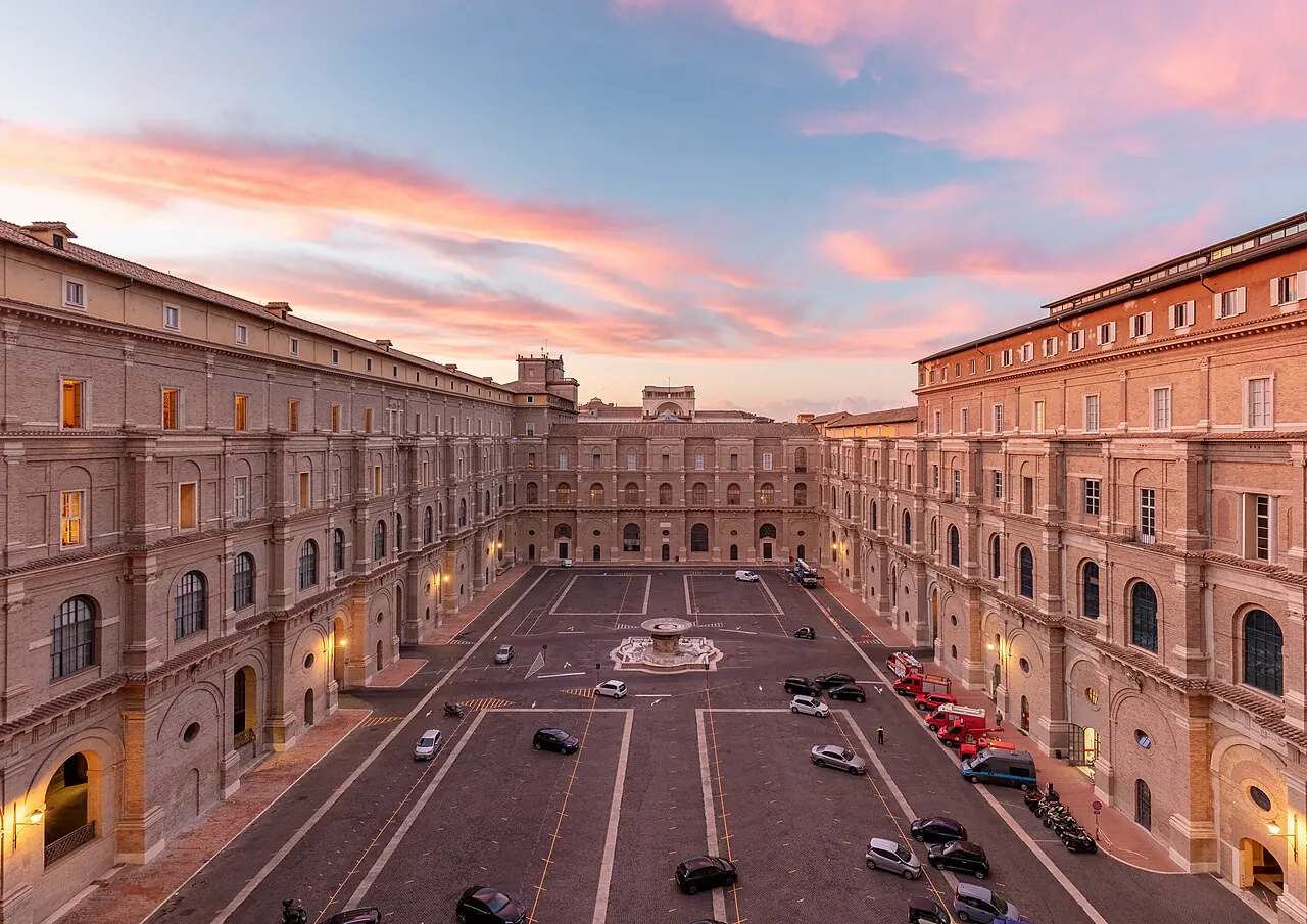 The Belvedere courtyard in Vatican City at sunset, seen from above one end: a long rectangular court walled in by four-storey Renaissance palace wings, its entire floor asphalt, parked nose to tail with cars, vans and coaches around a small central fountain.