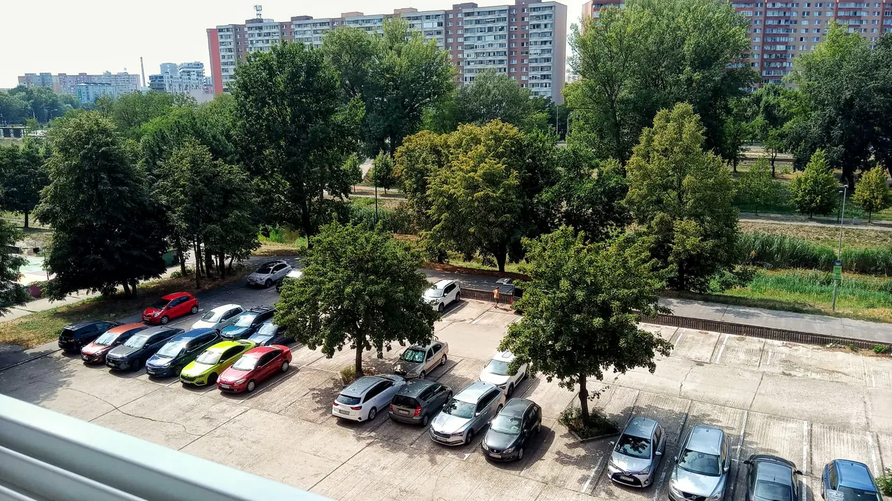 The view from a fifth-floor balcony in Petržalka, a white balcony rail across the bottom left corner: a wide concrete parking lot filling the foreground with two ranks of parked cars, a line of mature trees behind it, and a wall of long panel blocks along the horizon.