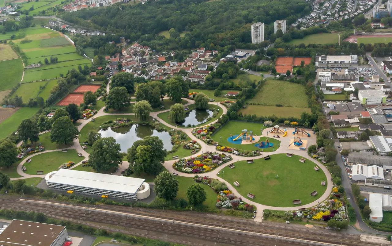 The same ground from the same height, reimagined as a public park: two ponds, lawns and curving gravel paths, beds of yellow, red and white flowers, benches and a playground with slides — the same town, the same fields, the same industrial estate, the same white multi-storey deck and the same railway line still framing it.