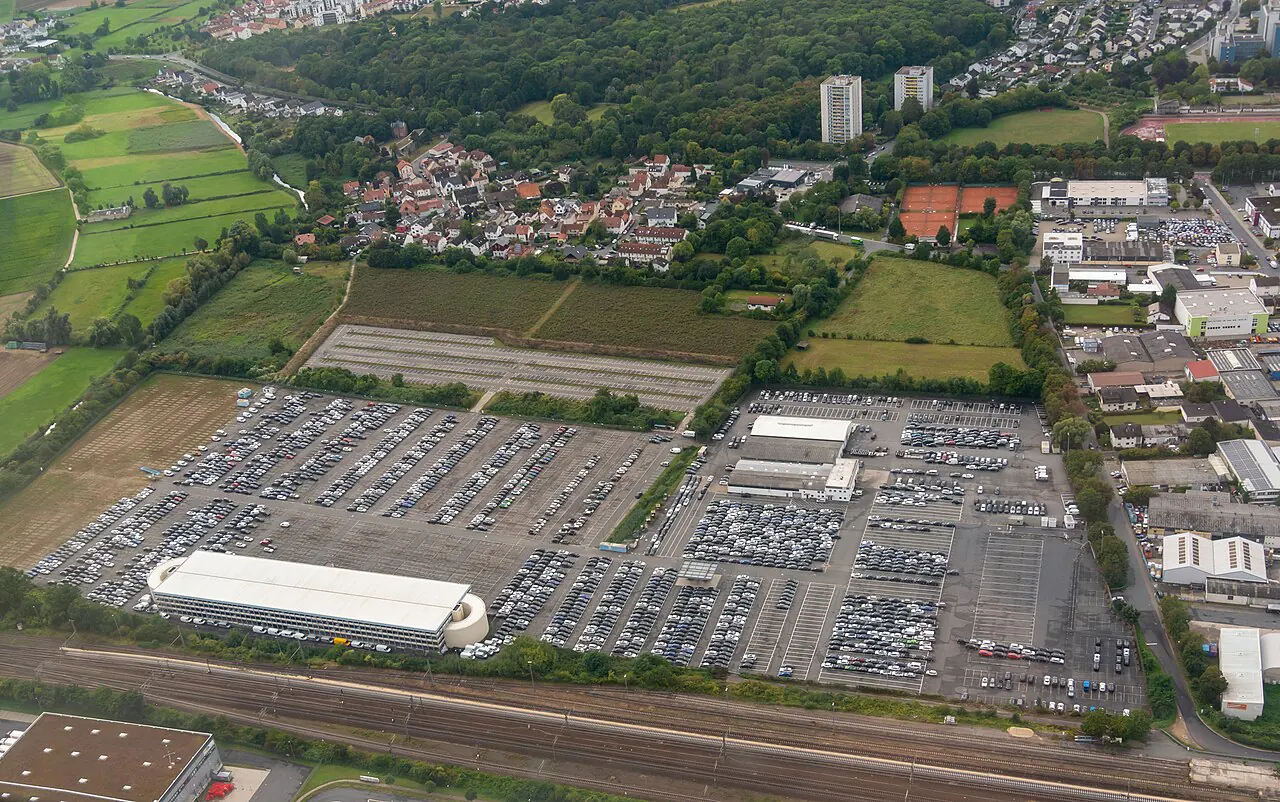 An aerial view of a car park on the edge of Gross-Gerau: several hectares of asphalt ruled into rows and packed end to end with new cars, a long white multi-storey deck at the lower left, ploughed and green fields to the left, the town — houses, two tower blocks and red tennis courts — along the top, an industrial estate down the right side and a multi-track railway line running across the bottom.